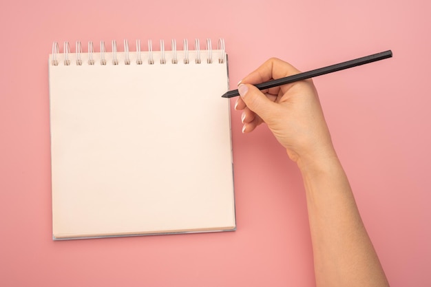 Mano de mujer de bloc de notas de maqueta con lápiz junto a un bloc de notas blanco en blanco vacío vista superior de la mano que sostiene el lápiz contra el bloc de notas en espiral sobre fondo blanco tomando notas concepto