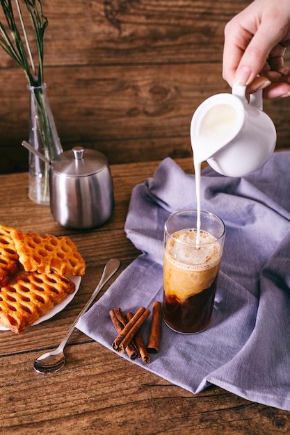 Mano de mujer con crema vertiendo leche en vaso con palitos de canela de café galletas caseras y bunc