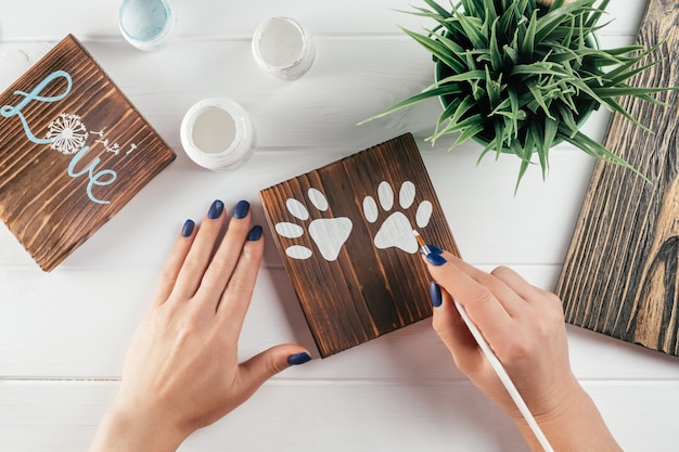 Mano de mujer dibuja huellas de patas en tablero decorativo de madera quemada con pincel de pintura blanca, bricolaje creativo