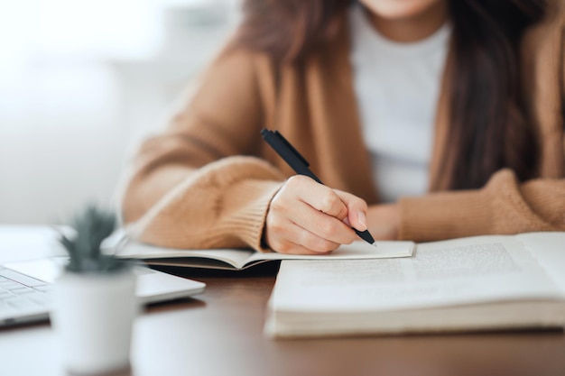 Mano de mujer escribiendo en un bloc de notas en espiral colocado en un escritorio de madera con varios artículos en casa