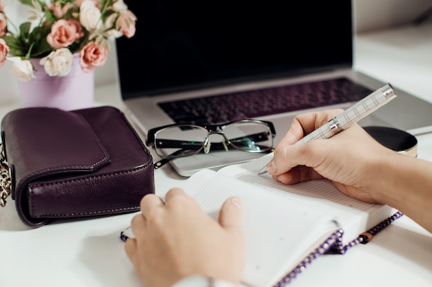 Mano de mujer escribiendo en el bloc de notas vacío colocado en el escritorio de la oficina con ordenador portátil, gafas, maceta de flores