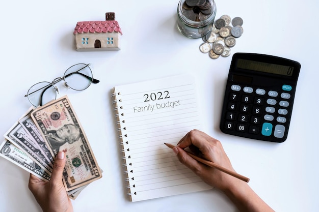 Mano de mujer escribiendo en el cuaderno mientras sostiene dinero y calculadora de su lado. Copie el espacio, vista superior. Concepto de presupuesto para el hogar.