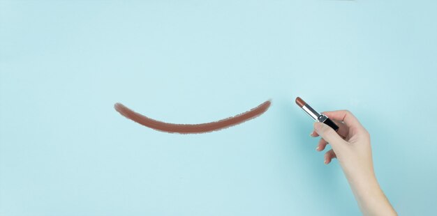 Mano de mujer escribiendo sonrisa con lápiz labial marrón sobre fondo azul.