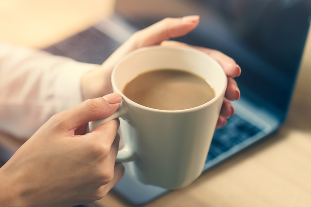 Mano de mujer en el escritorio con una computadora portátil, una taza de café, con la luz del sol de la mañana.