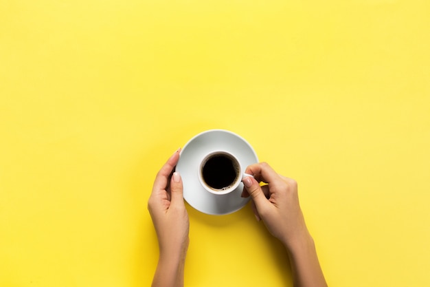 Mano de la mujer del estilo de Minimalistic que sostiene una taza de café en fondo amarillo. Colocación plana, vista superior