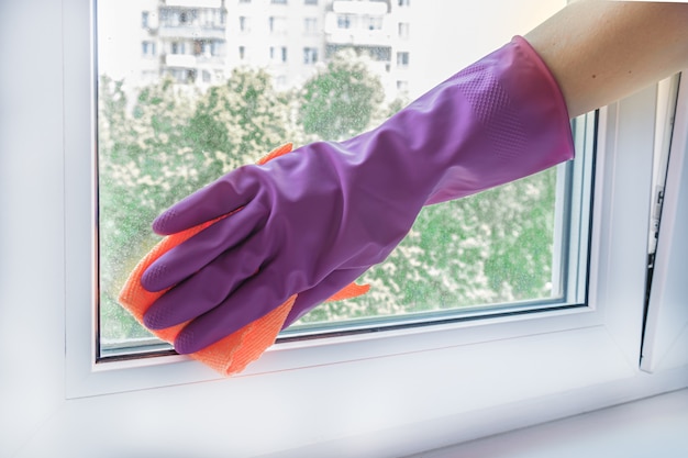 La mano de la mujer en un guante de goma lila limpia una ventana de la unidad de vidrio en una habitación con un trapo