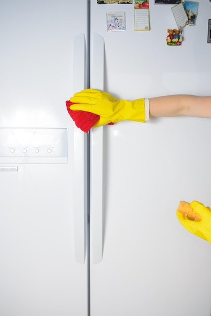 Una mano de mujer con guante protector de goma amarilla limpiando la puerta blanca del refrigerador cerrado con esponja naranja