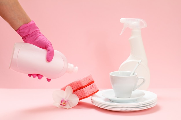 Mano de mujer en un guante protector de nitrilo con una botella de líquido para lavar platos, platos blancos, un platillo, una taza sobre el fondo rosa. Concepto de lavado y limpieza.