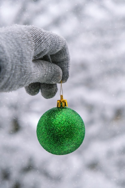 Foto la mano de la mujer en los guantes sostiene la bola de navidad verde brillante de moda en los abetos de las ramas nevadas en el bosque de invierno