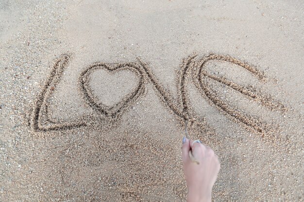 Foto mano de mujer haciendo el símbolo del corazón y la palabra amor escrita a mano en la playa de arena