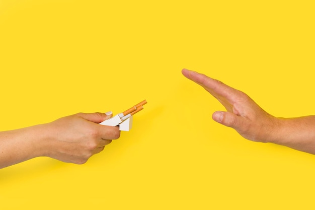Mano de mujer invitando a cigarrillos y mano de hombre con gesto de parada en un fondo amarillo con espacio de copia