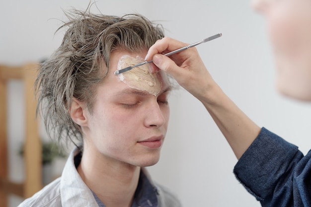 Mano de mujer joven con herramienta de belleza aplicando base en la cara del empresario contemporáneo en ropa casual en la oficina antes del trabajo