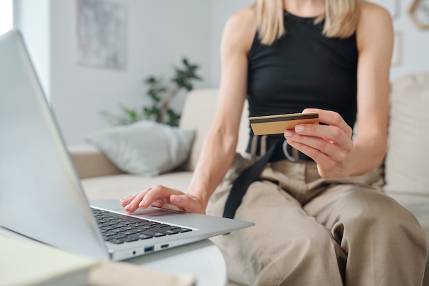 Mano de mujer joven en ropa casual con tarjeta de crédito sobre el teclado de la computadora portátil y presionando el botón mientras navega en la tienda en línea