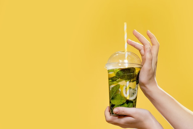 Mano de mujer joven sosteniendo una copa de cóctel con menta. Foto de estudio con fondo amarillo.