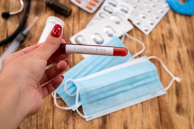 Mano de una mujer joven sosteniendo un tubo de ensayo con sangre sobre un fondo de medicamentos y máscaras. Concepto de medicina