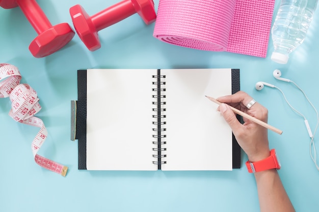 Mano de mujer con lápiz sobre cuaderno en blanco con deporte y equipos de Yoga en color pastel