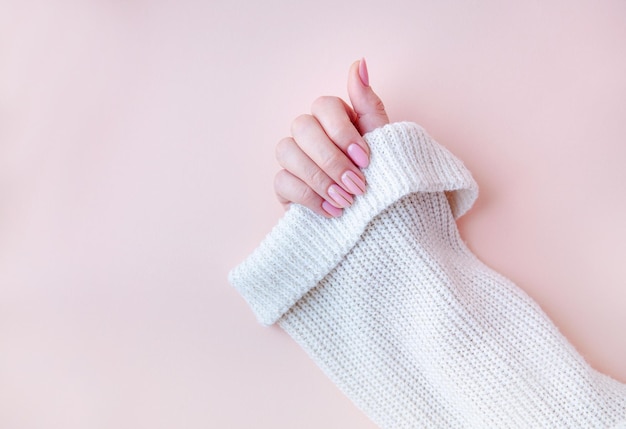 Una mano de mujer con una manicura en un suéter blanco sobre un fondo rosa Tono de esmalte de gel desnudo