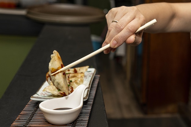 Mano de mujer con palillos sumergiendo una gyoza en salsa de soya y luego engulliéndola con deleite