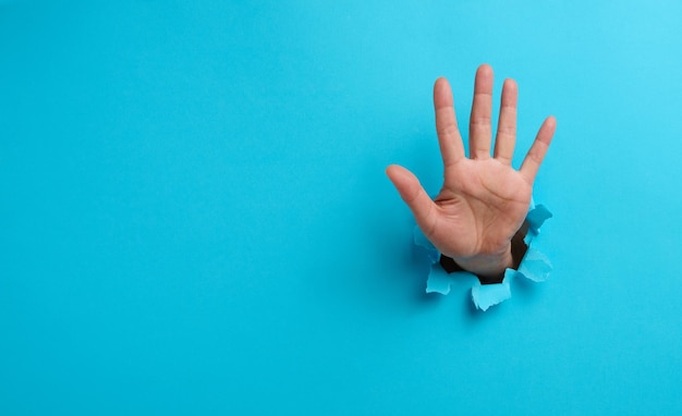 La mano de una mujer con la palma abierta sobresale de un agujero rasgado en papel azul, haciendo un gesto para detenerse