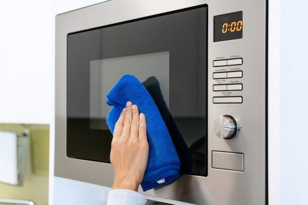 Mano de mujer con paño de microfibra limpiando la superficie del horno microondas. Hogar