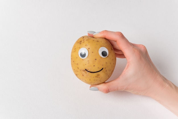 Mano de mujer con patatas crudas con cara divertida. Patatas con ojos saltones y sonrisa sobre fondo blanco.