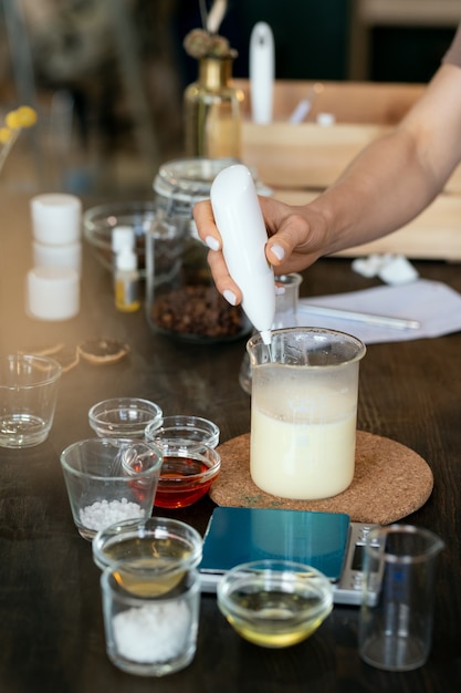 Mano de mujer poniendo sustancia líquida transparente en cristalería con masa de jabón derretido mientras hace la mezcla para cosméticos hechos a mano