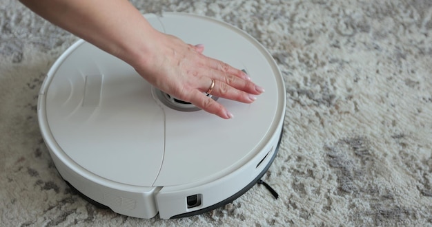 La mano de la mujer presiona el botón para encender el robot aspirador moderno Aspirador robótico en la sala de estar Limpieza del hogar