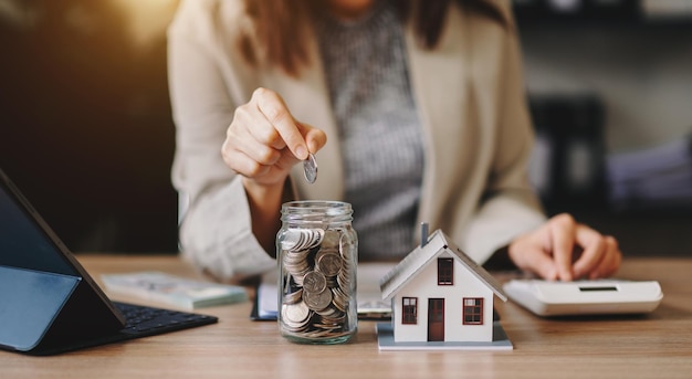 Mano de mujer puso monedas de dinero en una casa de madera sobre la mesa para ahorrar dinero para comprar una casa concepto financiero de ahorrar dinero para comprar una casa