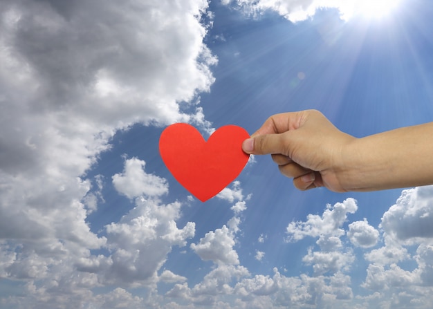 La mano de la mujer que lleva a cabo un corazón rojo en fondo del cielo.