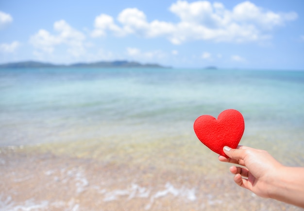 Mano de la mujer que lleva a cabo un corazón rojo en la playa con el fondo borroso del mar y del cielo azul. Concepto de amor