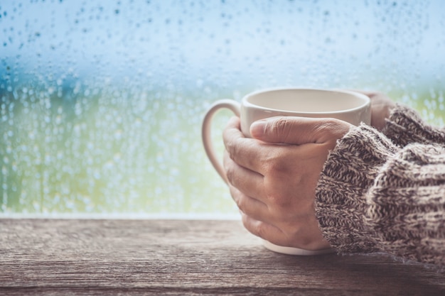 Mano de la mujer que sostiene la taza de café o té en el fondo de la ventana del día lluvioso