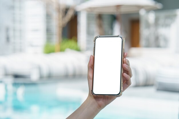 Mano de la mujer que sostiene el teléfono móvil de la pantalla blanca en blanco cerca de la piscina.