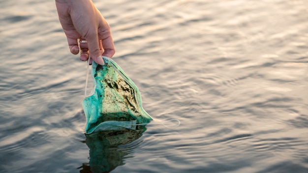 Foto mano de mujer recogiendo una máscara médica desechable usada desechada flota en aguas marinas. residuos plásticos de coronavirus que contaminan el medio ambiente. basura en la playa que amenaza la salud de los océanos.