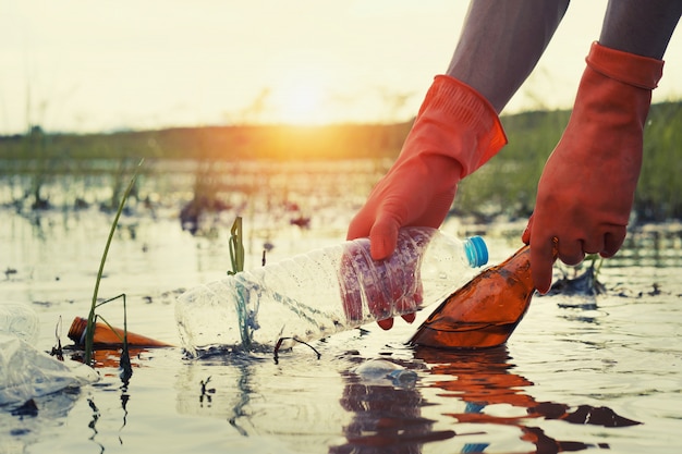 Mano de mujer recogiendo plástico de basura para limpiar en el río con puesta de sol