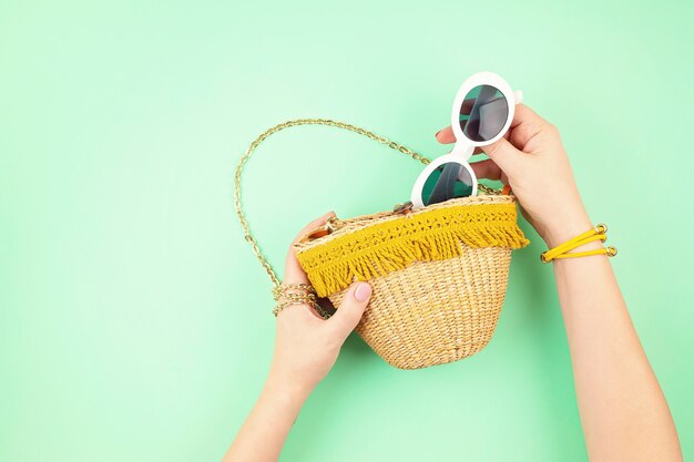 Mano de mujer sosteniendo una bolsa de paja con accesorios de vacaciones de verano. Vacaciones de verano, viajes a países tropicales, mar, concepto de estilo de verano. Vista superior, endecha plana