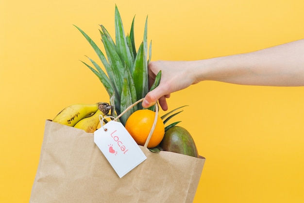 Mano de mujer sosteniendo una bolsa de papel llena de frutas tropicales sobre fondo amarillo. Copie el espacio.