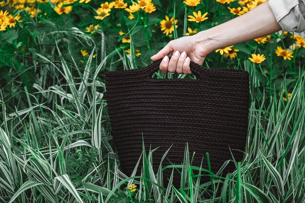 Mano de mujer sosteniendo una bolsa de punto marrón sobre un fondo de hierba y flores