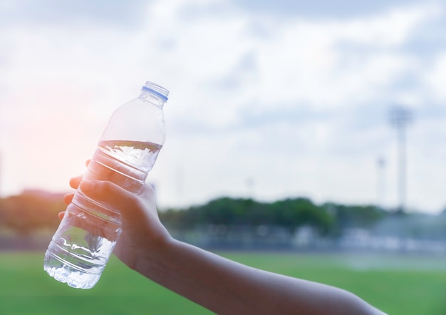 Mano de mujer sosteniendo una botella de agua potable en cielo azul y campo verde