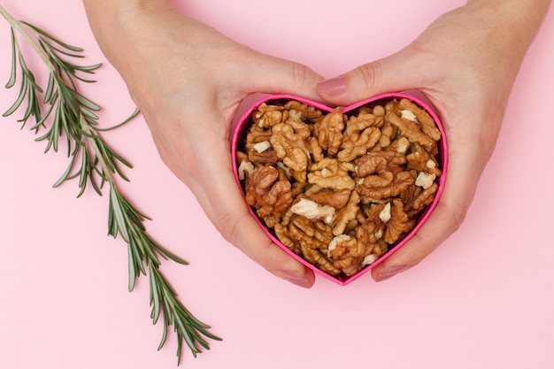 Mano de mujer sosteniendo una caja en forma de corazón con nueces peladas sobre fondo rosa.