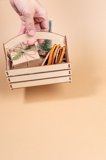 Mano de mujer sosteniendo una caja de madera con Navidad - árbol de Navidad decorativo de adornos, rodajas de naranja secas, caja de regalo envuelta de bricolaje sobre fondo beige. Imagen navideña. Orientación vertical. Copie el espacio.