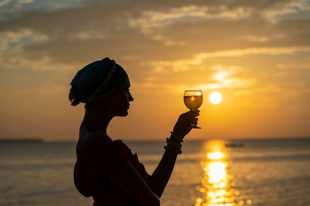 Mano de mujer sosteniendo una copa de vino contra una hermosa puesta de sol cerca del mar en la playa tropical de cerca