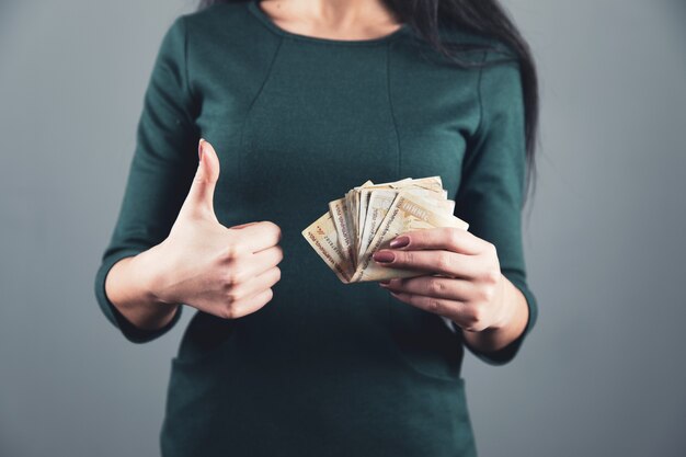 Mano de mujer sosteniendo dinero y signo bien sobre fondo gris