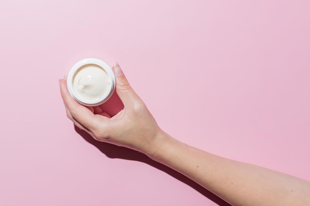 Mano de mujer sosteniendo un recipiente de crema cosmética abierto sobre fondo rosa Maqueta con espacio para texto Presentación de marca de producto cosmético