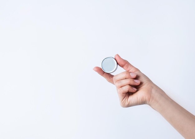 Mano de mujer sosteniendo un recipiente de estaño redondo en blanco para bálsamo labial o crema sobre fondo gris claro