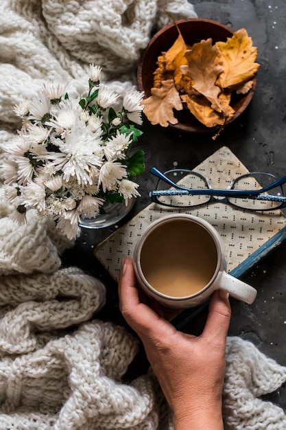 Mano de mujer sosteniendo taza de café libro acogedoras hojas de otoño a cuadros sobre fondo oscuro vista superior