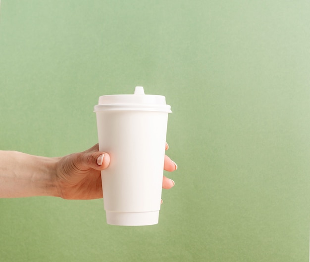 Mano de mujer sosteniendo una taza de café de papel para llevar grande blanca maqueta sobre fondo verde