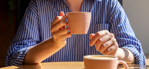 Mano de mujer sosteniendo la taza de café sobre la mesa de madera en el café en un día soleado con luz diurna closeup