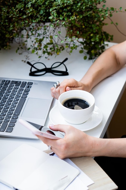 Mano de mujer sosteniendo una taza de café y un teléfono inteligente en una oficina acogedora con computadora portátil y plantas verdes