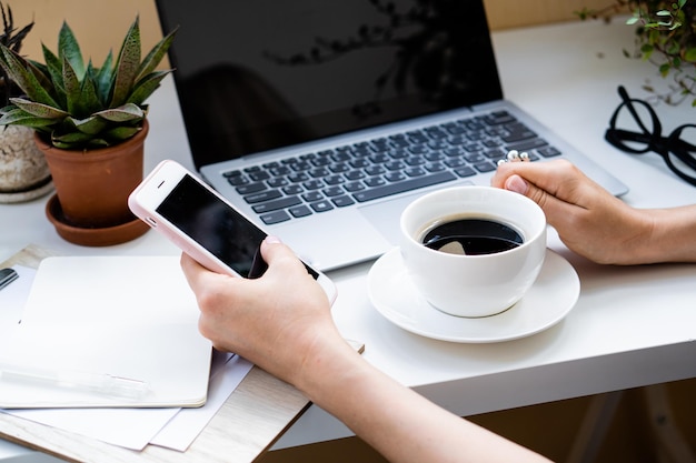 Mano de mujer sosteniendo una taza de café y un teléfono inteligente en una oficina acogedora con computadora portátil y plantas verdes