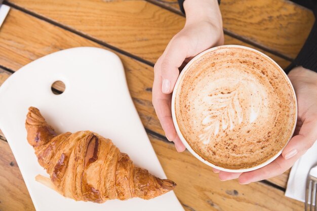 Mano de mujer sosteniendo una taza grande con café capuchino y croissant francés recién hecho en una mesa de madera